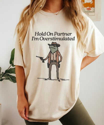 Cream t-shirt with “Hold On Partner I’m Overstimulated” text and a small cowboy character graphic
