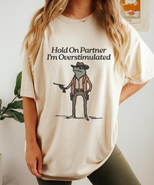 Cream t-shirt with “Hold On Partner I’m Overstimulated” text and a small cowboy character graphic